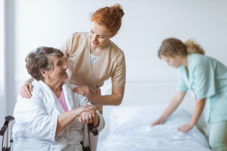 Elderly woman on wheelchair in nursing home with helpful doctor at her side and young nurse making the bed