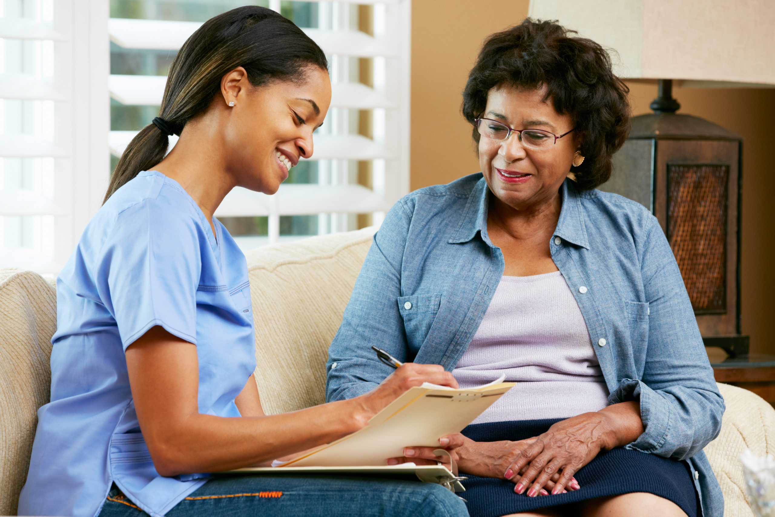 A nurse in a blue uniform sits on a couch and smiles while taking notes on a clipboard. She is interacting with an elderly woman wearing glasses, a blue denim shirt, and a light purple top, who is also seated and smiling. They appear to be in a comfortable living room.