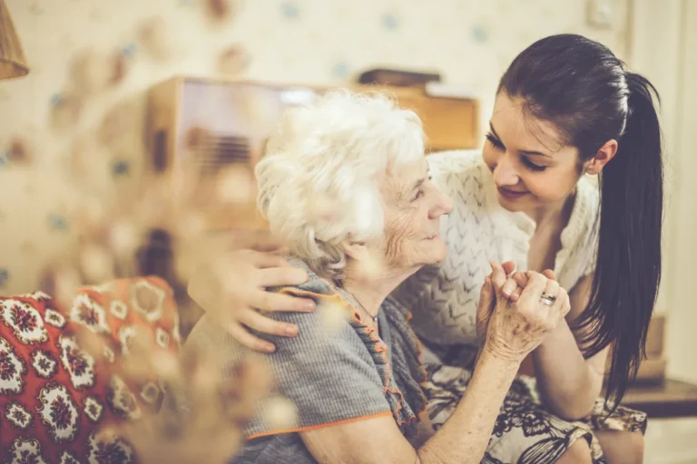 A young woman with dark hair smiles and embraces an elderly woman with white hair. The elderly woman is seated and wearing a gray outfit, while the young woman is standing close beside her, wearing a light-colored top. They are holding hands and looking at each other affectionately.