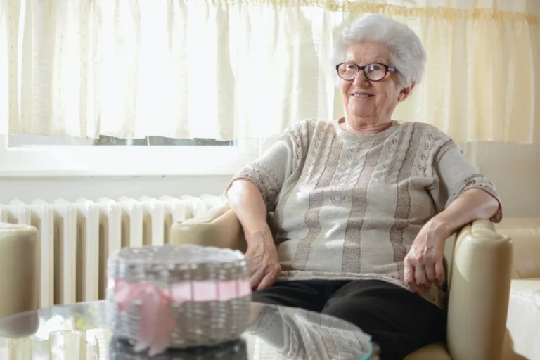An elderly woman with short white hair and glasses is sitting on a beige armchair. She is smiling and wearing a knitted sweater. A small, partially visible, white basket with pink ribbon sits on a glass table in front of her. The background has light-colored curtains.