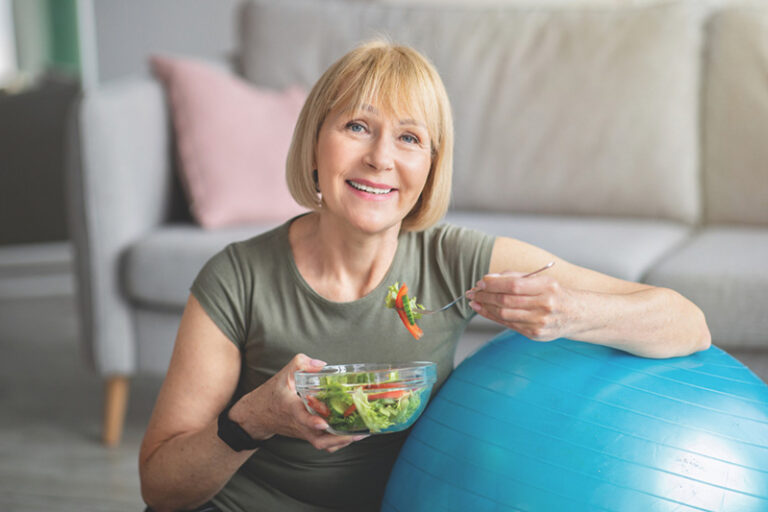 Smiling woman with short blond hair sits indoors near a blue exercise ball, holding a bowl of salad and a fork. A gray sofa with a pink cushion is in the background.