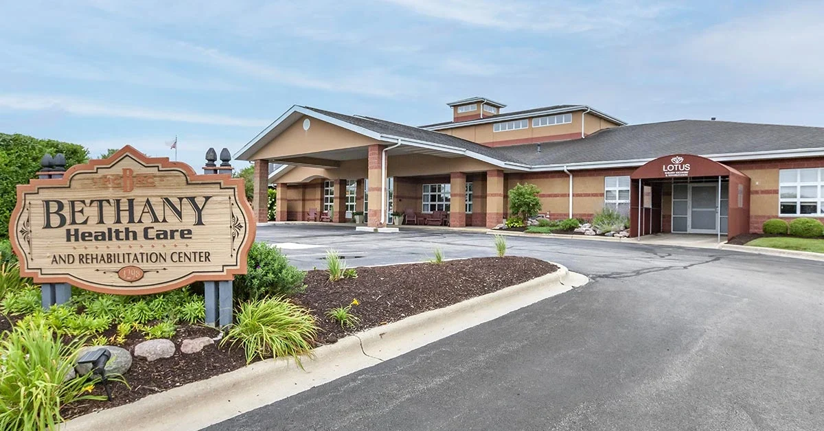 An exterior view of Bethany Rehabilitation & Health Care Center. The building has a covered entrance with paved walkways and landscaping featuring green plants. A sign in the foreground reads 