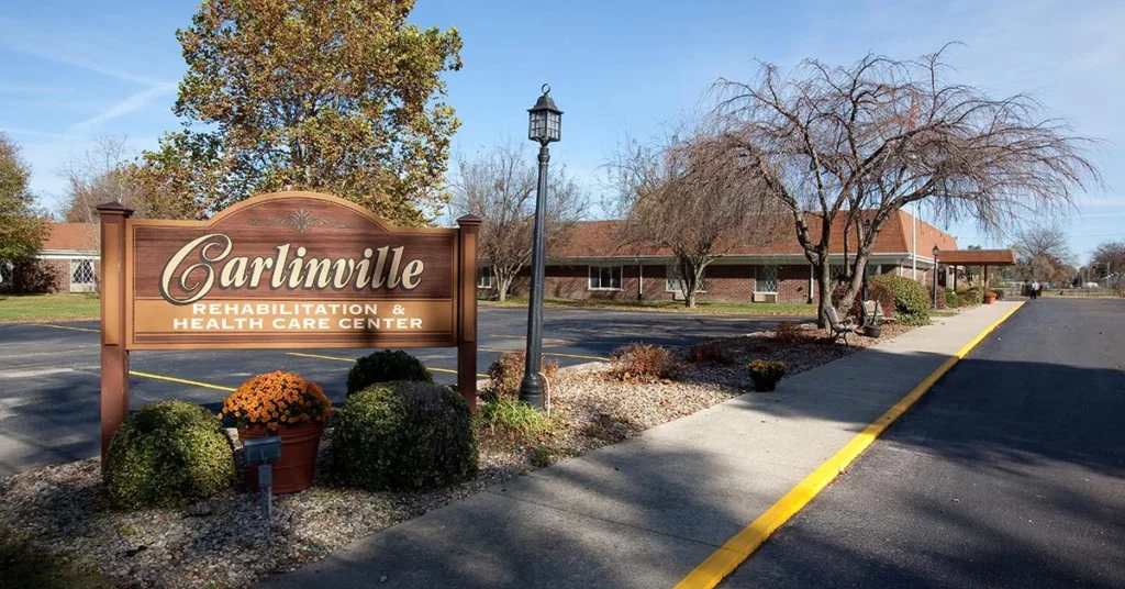 A sign for Carlinville Rehabilitation & Health Care Center stands near a landscaped area with mulch and orange flowers. In the background, the one-story brick building with a red roof and lamp post beside a paved walkway adds to the welcoming ambiance of the Rehabilitation & Health Care Center.