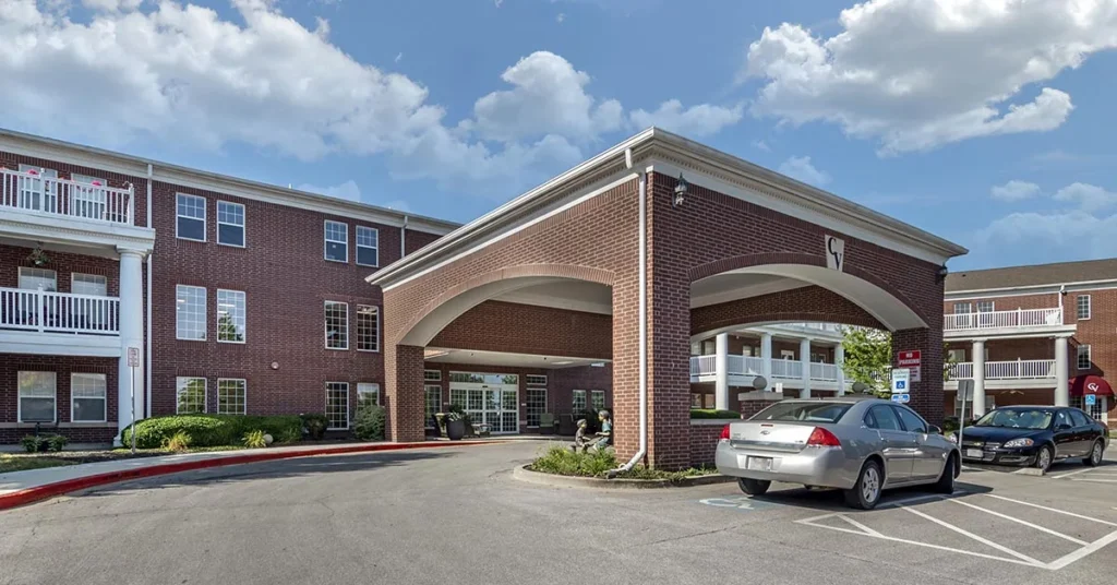 A three-story brick building with white trim in this Senior Living Community features a portico at the entrance, supported by brick columns. A silver car is parked near the entrance. Balconies and windows are visible on the upper floors, and the sky is clear with a few clouds.