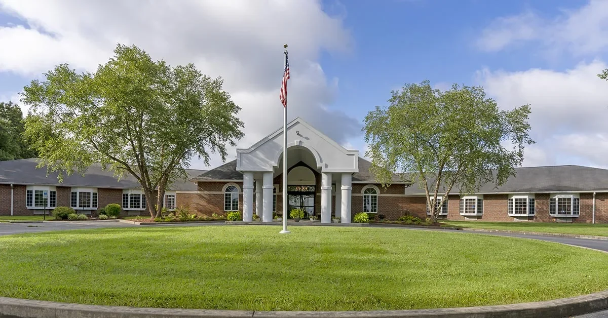 A one-story brick Rehabilitation & Health Care Center with white pillars at the entrance is surrounded by green lawns and trees. An American flag on a flagpole stands prominently in front of the building, with a circular driveway framing the grassy area. The sky is partly cloudy.