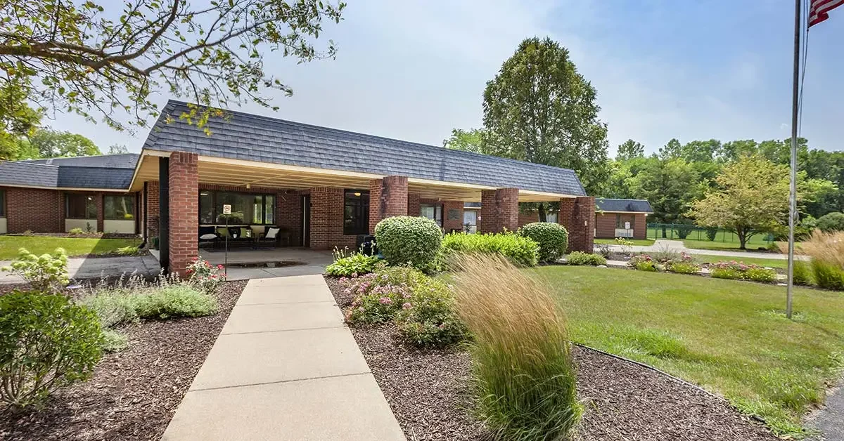 A single-story Rehabilitation & Health Care Center with a brick facade and dark shingle roof, surrounded by neatly landscaped gardens and shrubs. A concrete walkway leads to the entrance, and a flagpole is visible on the right. Trees and a grassy area are in the background under a clear sky.