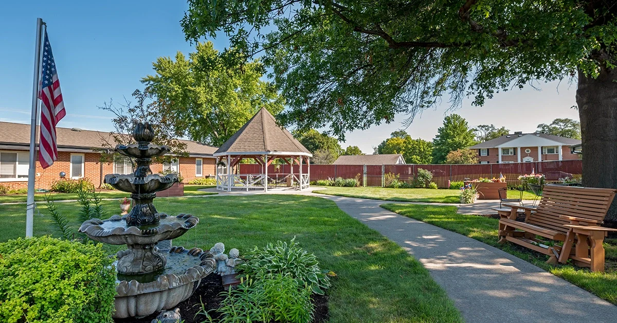 A tranquil garden at the Rehabilitation & Health Care Center features a paved pathway leading to a gazebo surrounded by greenery. An American flag stands beside a tiered water fountain. Benches sit on the right near a large tree, with a building and white fence in the background.