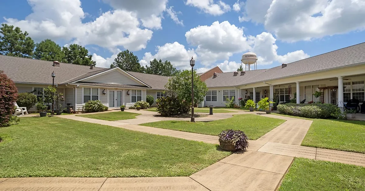 A well-maintained courtyard in this assisted living community is surrounded by single-story buildings with gray siding and white trim. The courtyard features lush green grass, several small bushes, walkways, and a few street lamps. A water tower and trees are visible in the background under a partly cloudy sky.