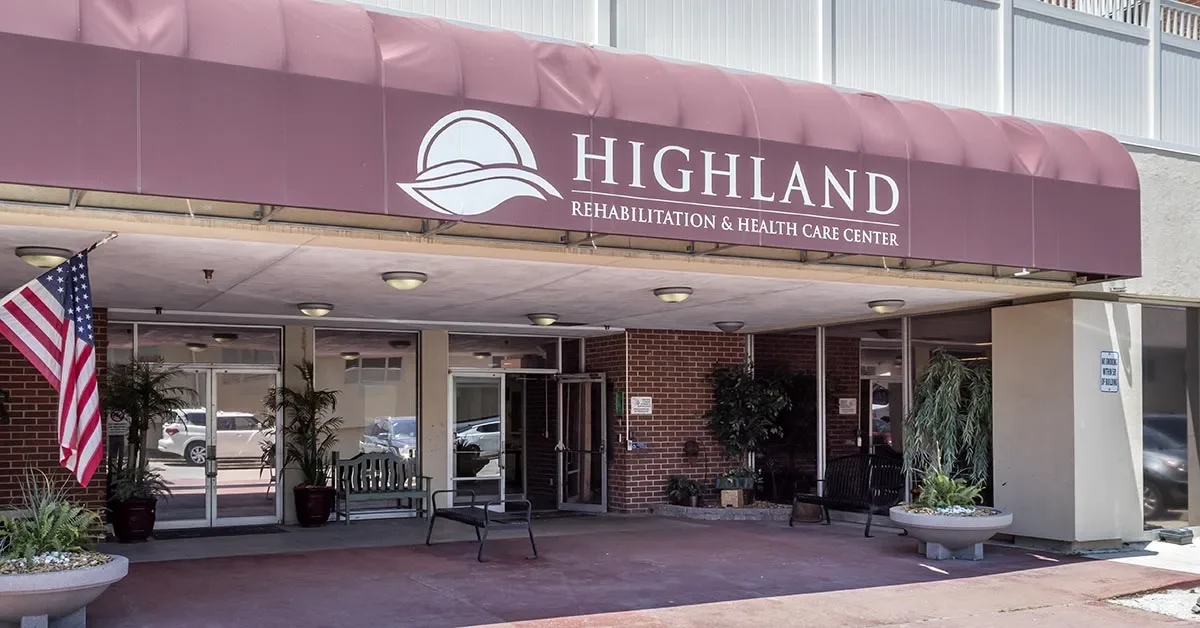 The entrance of Highland Rehabilitation & Health Care Center features a maroon awning with the center's name and logo. Potted plants, benches, an American flag, and a sign indicating parking create a warm welcome. The brick and glass exterior emphasizes its accessible design.