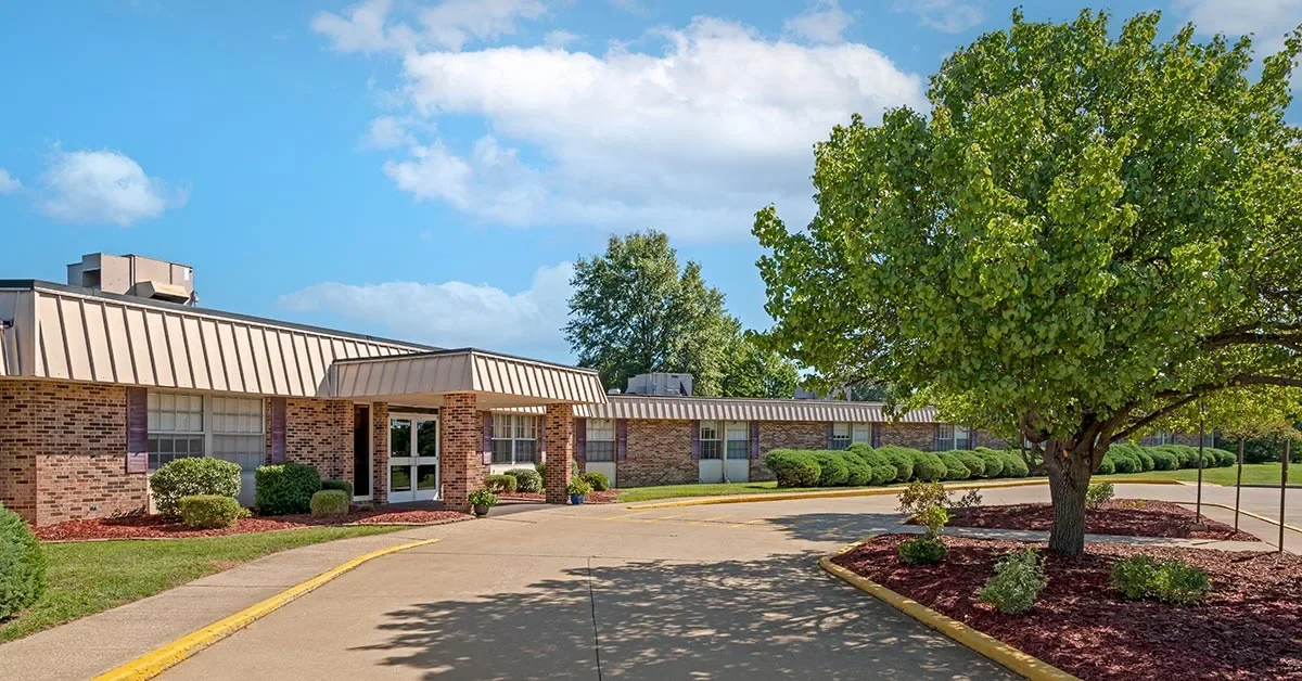 A one-story Rehabilitation & Health Care Center with a brick exterior is surrounded by well-maintained landscaping, including bushes and a large leafy tree. A paved walkway with curbs leads to the entrance under a blue sky dotted with clouds.