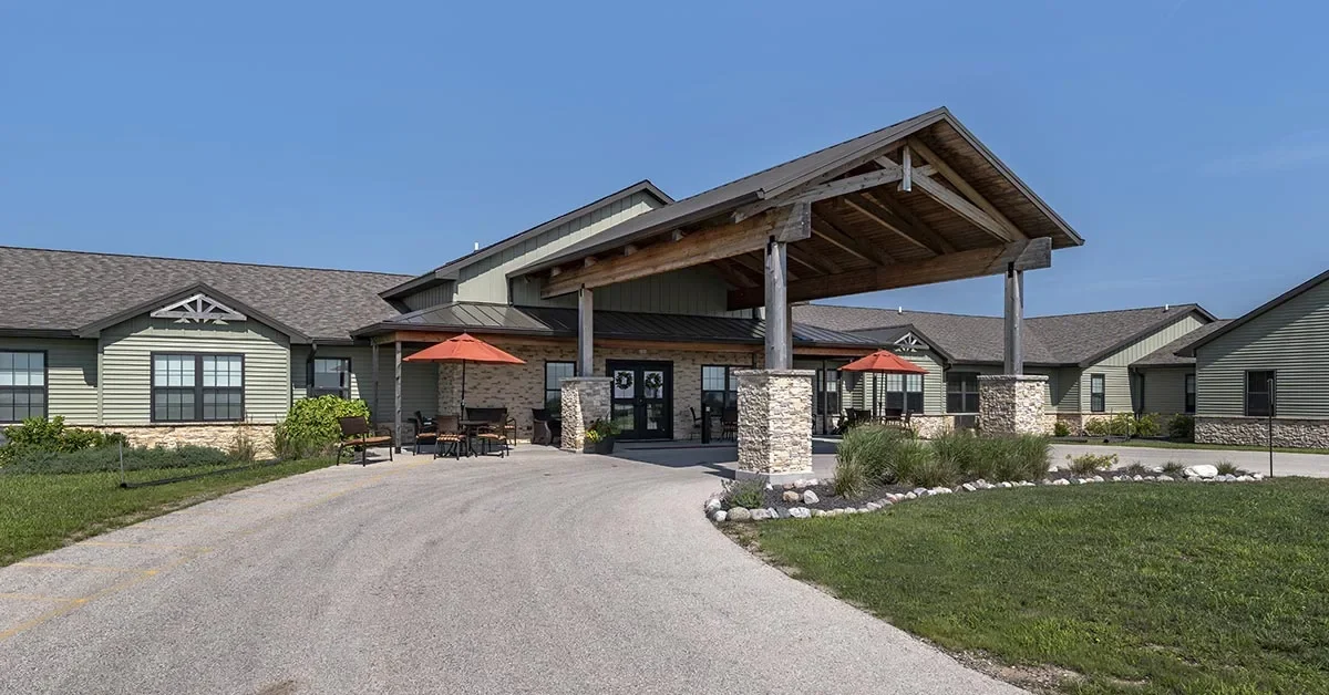 Exterior of a single-story Assisted Living & Memory Care building with green siding and stone accents, featuring a gabled roof and a covered entrance. The driveway leads to the entrance, with outdoor seating and red umbrellas visible on the left side under clear blue skies.