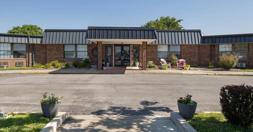 A single-story brick building with large windows and a central entrance under an awning welcomes visitors to the Rehabilitation & Health Care Center. The entrance features glass doors flanked by potted plants and outdoor seating. The foreground shows a paved driveway and more potted plants, with trees visible in the background.