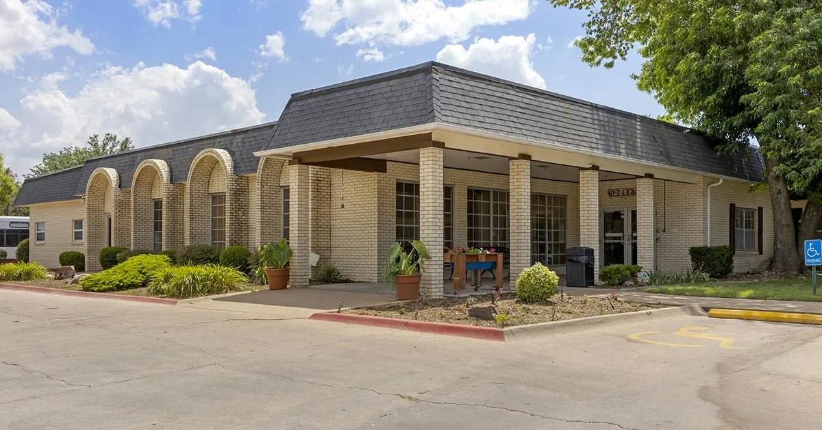 A one-story building with a flat roof and light-colored brick exterior, designed for senior rehabilitation. The entrance is covered by a small overhang supported by columns, and the building features large windows with landscaped plants along the front. A parking lot includes a designated accessible spot.