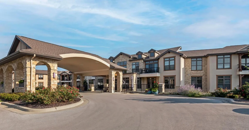 A two-story senior living building with beige walls and a gray roof, featuring a large covered entrance supported by stone pillars. The landscaping includes well-kept bushes and flower beds in front. The sky above is blue with scattered clouds.