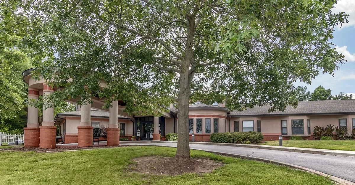 A Rehabilitation & Health Care Center, this single-story building features a rounded entrance, large front windows, and red brick pillars under a tree with lush green foliage. A driveway curves in front, encircled by verdant grass and vegetation. The sky is partly cloudy.