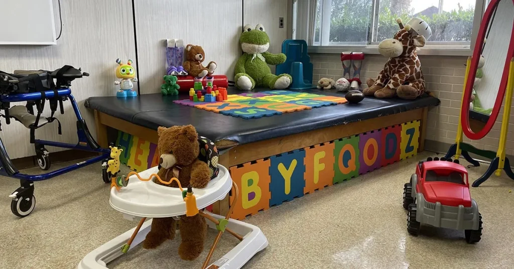 A children's playroom with an assortment of toys at the Specialty Center. A stuffed bear in a walker is in the foreground. A low table with colorful letters on the side holds more toys, including stuffed animals, blocks, and a frog plush. A walker and red toy truck also adorn the space.
