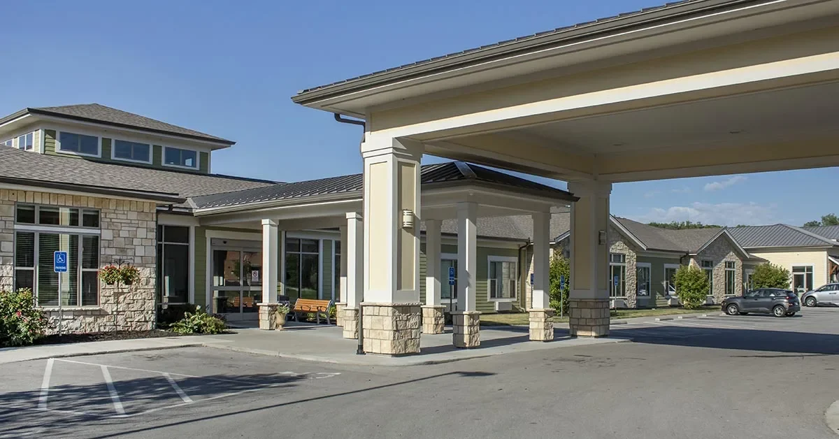 A modern Rehabilitation & Health Care Center housed in a single-story building with a stone and siding exterior, featuring a covered drop-off area supported by columns. The facility has multiple windows, manicured landscaping, and a parking lot with designated spaces. The sky is clear and sunny.