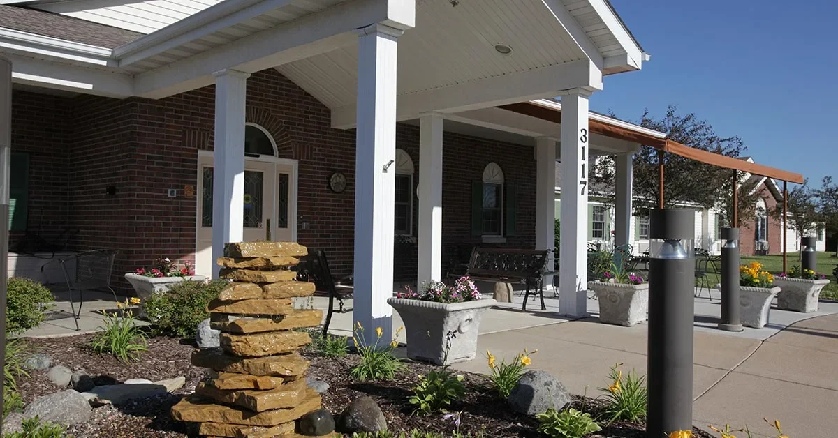 A building entrance with white pillars and a covered roof. There is a stack of rocks in the foreground among landscaped plants and flowers. Benches, potted plants, and a sidewalk surround the assisted living & memory care entrance. The building has brick walls and arched windows.