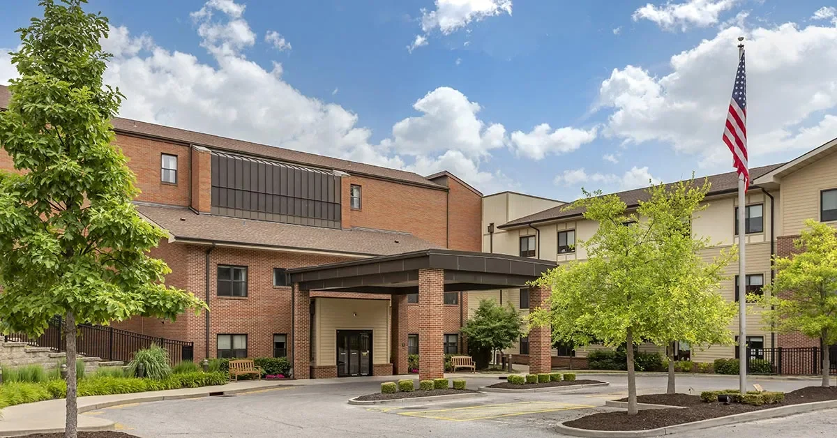 A modern multi-story building with a combination of brick and beige siding. The entrance features a covered drive-up area. The landscape includes green trees and manicured bushes. An American flag is on a tall pole in the foreground, and the sky is partly cloudy.