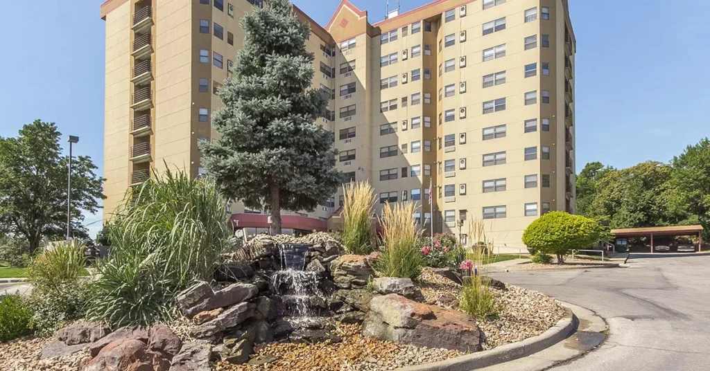 A tall beige apartment building with multiple windows is surrounded by a landscaped area, catering to 55+ apartments. In the foreground, a small waterfall flows over rocks, flanked by various plants and a tall pine tree. A driveway curves in front of the building.