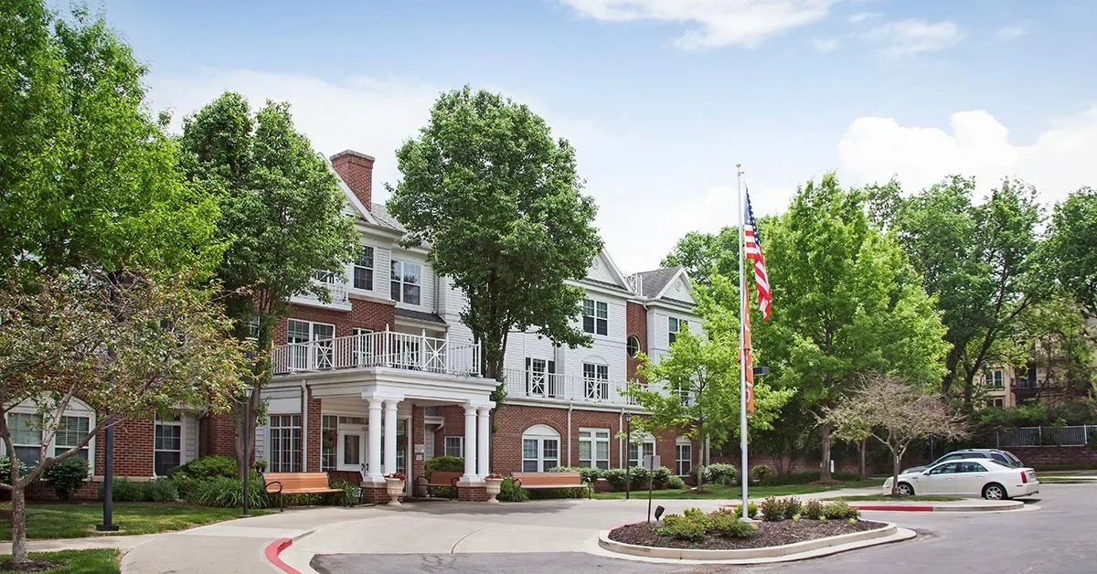 A three-story brick building with white accents, surrounded by lush trees and greenery, serves as a pleasant senior living facility. An American flag flies from a flagpole in the front yard. Several benches are placed near the entrance, and a paved driveway leads to a white car parked nearby.