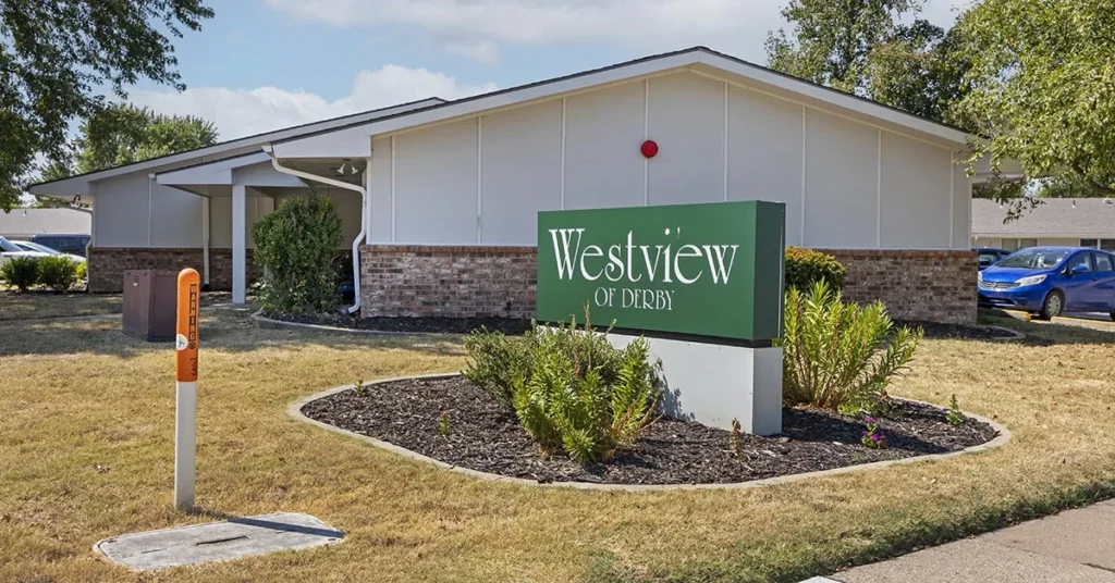 A single-story building with a beige exterior and brick detail is partially visible. In front, there's a green sign with white text reading 