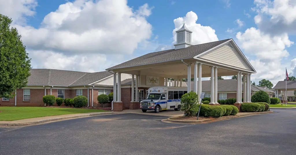 A Rehabilitation & Health Care Center housed in a one-story brick building with a white overhang and cupola. A small bus with blue and white accents is parked in front of the entrance. The surrounding area is landscaped with bushes and trees, and the sky is partly cloudy.
