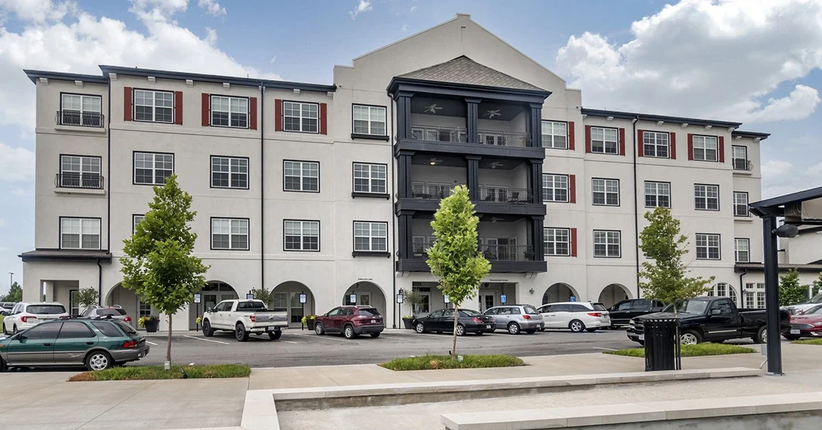 Four-story apartment building with a white and gray exterior, large windows, and balconies on the third floor. Several cars are parked in the lot in front of the building. Three small trees and a concrete area—perfect for rehabilitation and recovery—are visible under a partly cloudy sky.