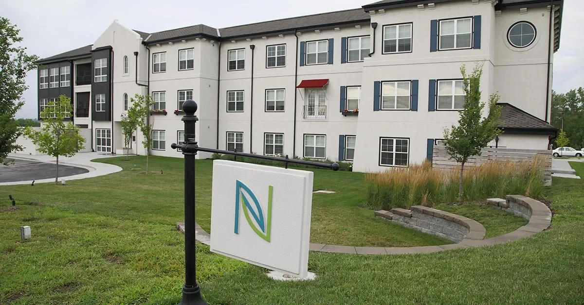 A modern three-story apartment building with white walls and dark window frames is surrounded by a well-kept lawn and landscaping. A sign with a stylized 