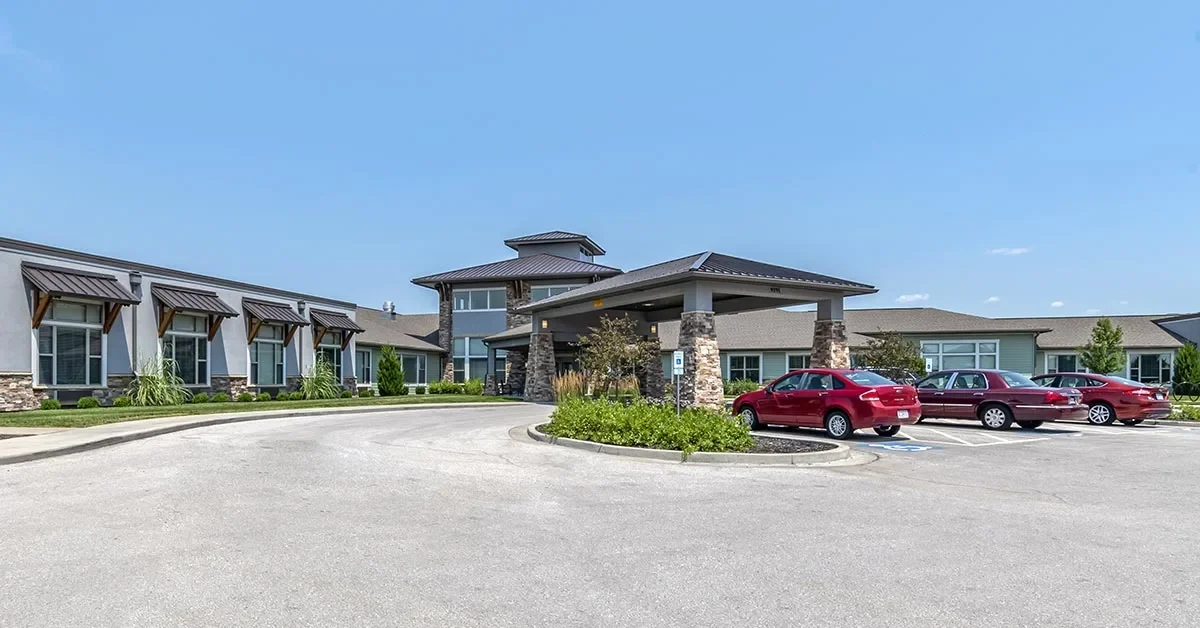 A modern, single-story building with a blend of stone and siding exterior, featuring a covered entrance. Several red cars are parked in a circular driveway in front. Under the clear blue sky, the area is surrounded by neatly manicured landscaping, making it an inviting Rehabilitation & Health Care facility.