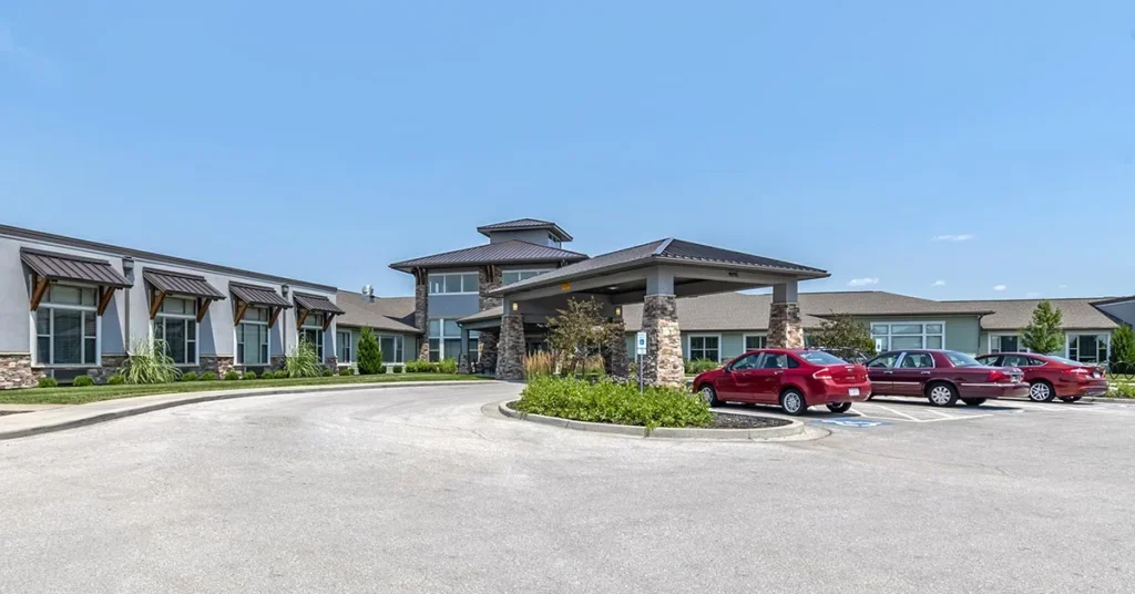 A modern, single-story building with a blend of stone and siding exterior, featuring a covered entrance. Several red cars are parked in a circular driveway in front. Under the clear blue sky, the area is surrounded by neatly manicured landscaping, making it an inviting Rehabilitation & Health Care facility.