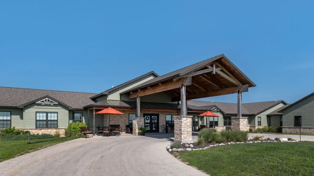 A modern, single-story building with a wooden and stone facade, featuring a large covered entrance with exposed beams. There are outdoor seating areas with red umbrellas, a curved driveway, and landscaped greenery against a clear blue sky.