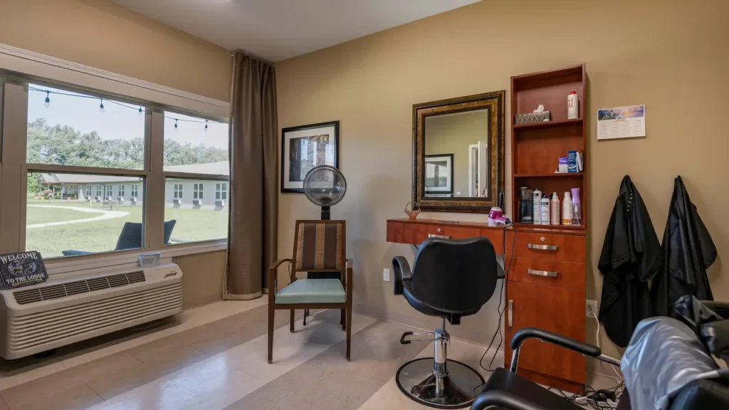 A cozy hair salon room with beige walls, featuring a styling chair in front of a large mirror on a wooden cabinet holding various hair products. To the left, a chair sits under a hair dryer near a window with a view of a green lawn outside. An air conditioner is beneath the window.