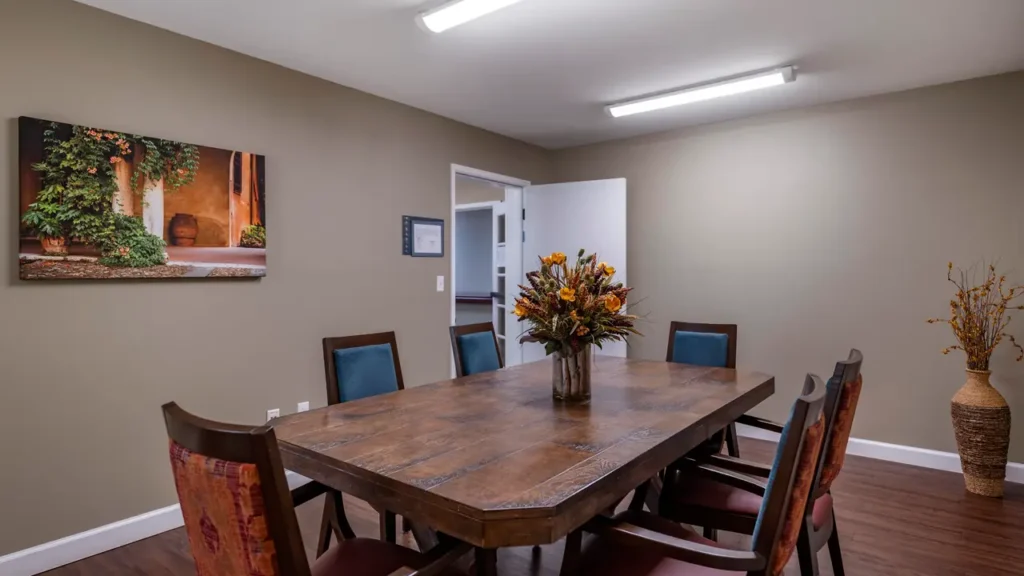 A meeting room with a large wooden table surrounded by eight chairs. A floral centerpiece sits on the table. The room has beige walls, wooden flooring, a painting on one wall, and an open door. Ceiling lights provide illumination.