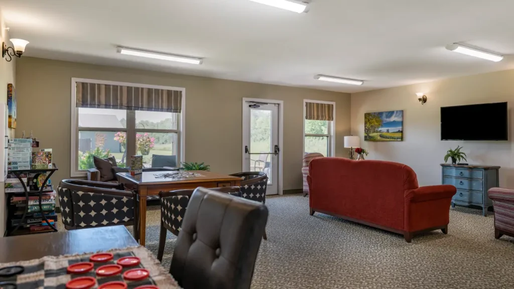 A cozy living room features a red sofa, a wall-mounted TV, and a nature-themed painting. There's a dining table with a plant centerpiece near large windows, and a gaming table with checkers in the foreground. Soft lighting and striped curtains add warmth to the space.