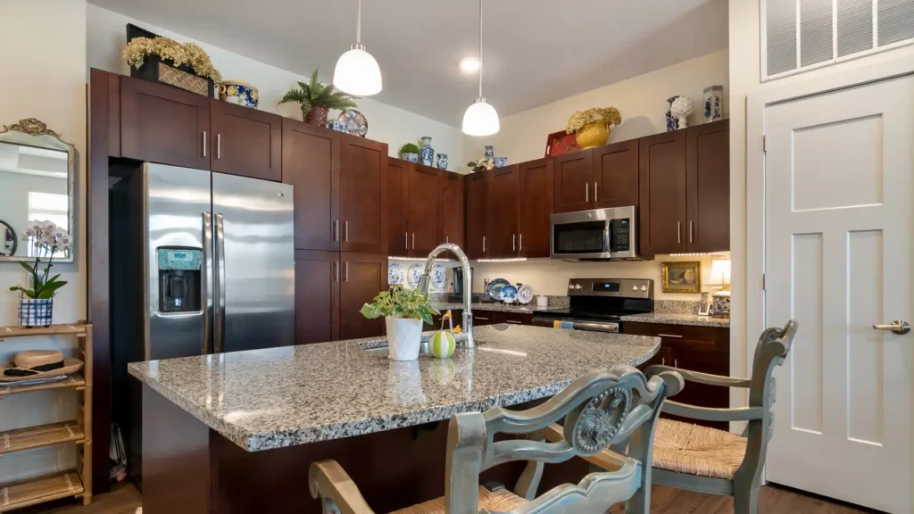 A modern kitchen with dark wood cabinets, stainless steel appliances, and a granite island countertop. There are pendant lights above the island, which features a sink and a small plant. Decorative plates and plants are displayed on top of the cabinets.