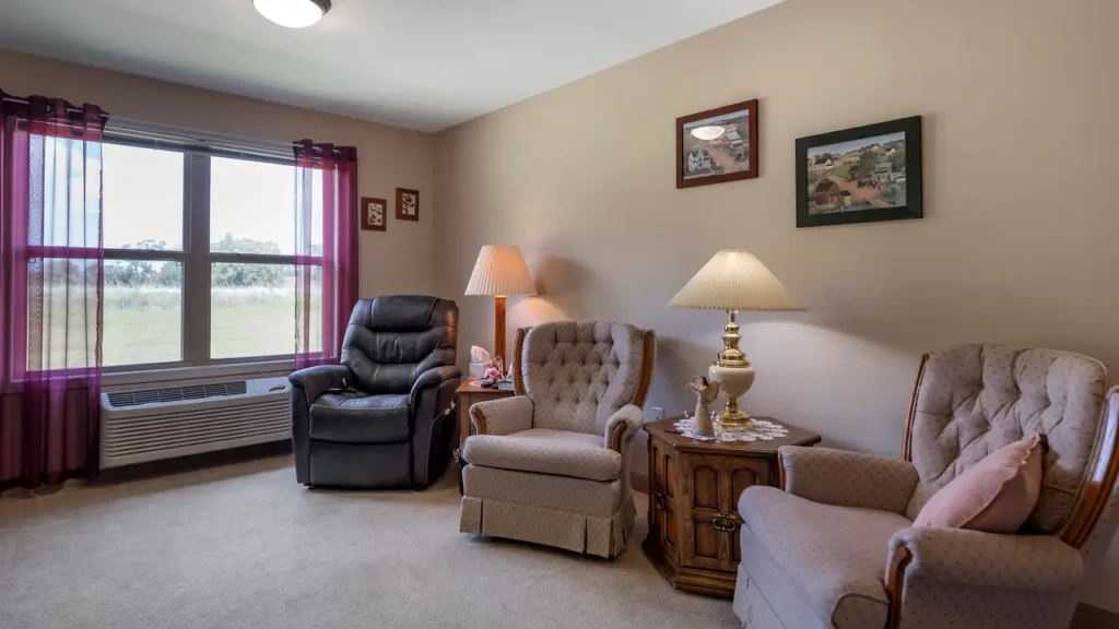 A cozy living room with three armchairs, two beige and one black. A wooden side table with two lamps is between the beige chairs. The room has beige walls with framed pictures and a large window with red curtains, allowing natural light to enter.