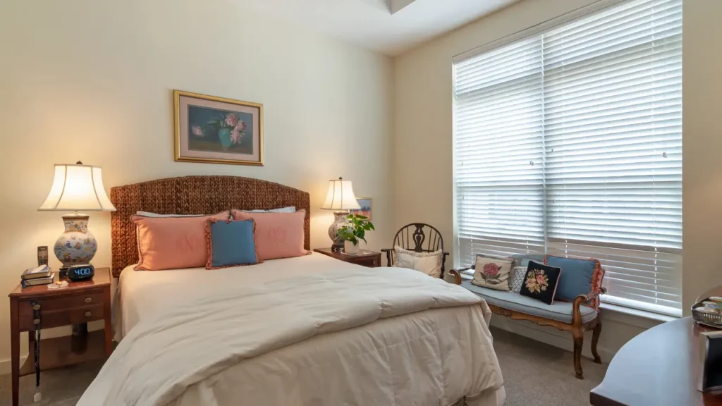 A cozy bedroom with a large bed featuring pink pillows and a blue cushion. Two side tables with lamps flank the bed. A bench with pillows sits by the window, which has blinds partially open, allowing natural light to fill the room. A framed artwork hangs above the bed.