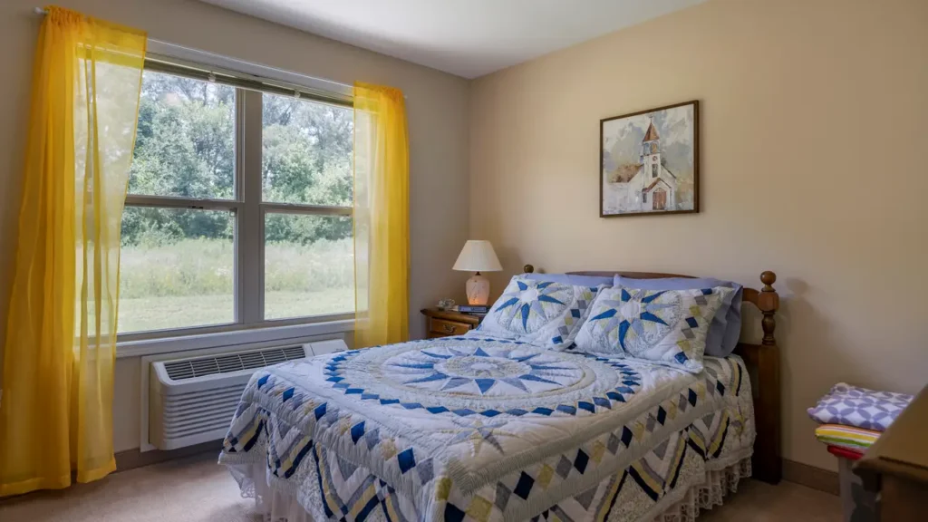 A small bedroom featuring a twin bed with a blue and yellow quilted bedspread. The room has beige walls, a window with yellow curtains, an AC unit below, and a nightstand with a lamp. A framed picture of a church hangs above the bed. The window offers a view of greenery.