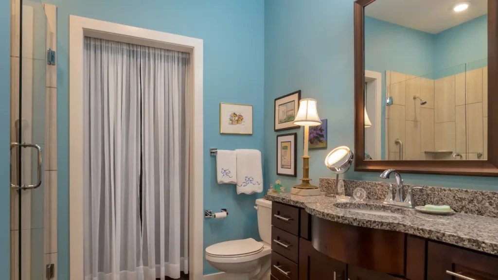 A bathroom with light blue walls features a large mirror above a granite countertop with a sink and a small lamp. A toilet is next to the vanity, with a towel rack above it holding two white hand towels. A glass door with a sheer curtain leads outside. Various framed pictures hang on the wall.