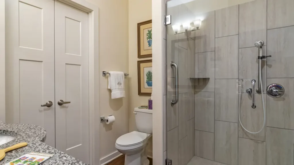 A bathroom with a glass-enclosed shower featuring beige tiles. There's a white toilet next to it with a white towel hanging on a rack above a toilet paper holder. Two framed pictures are on the wall behind the toilet. A door is on the left.