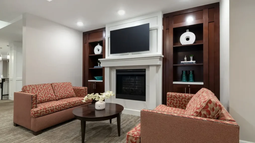 A cozy living room features a central white fireplace with a mounted flat-screen TV above it. Two patterned orange-red sofas surround a round coffee table adorned with a white floral arrangement. Built-in wooden shelves with decorative objects frame the fireplace.