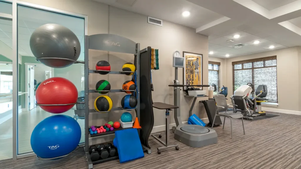 A fitness room with various gym equipment, including exercise balls, foam rollers, medicine balls, hand weights, a balance trainer, a stationary bike, and an elliptical machine. The room has large windows and modern lighting. A motivational poster is on the wall.