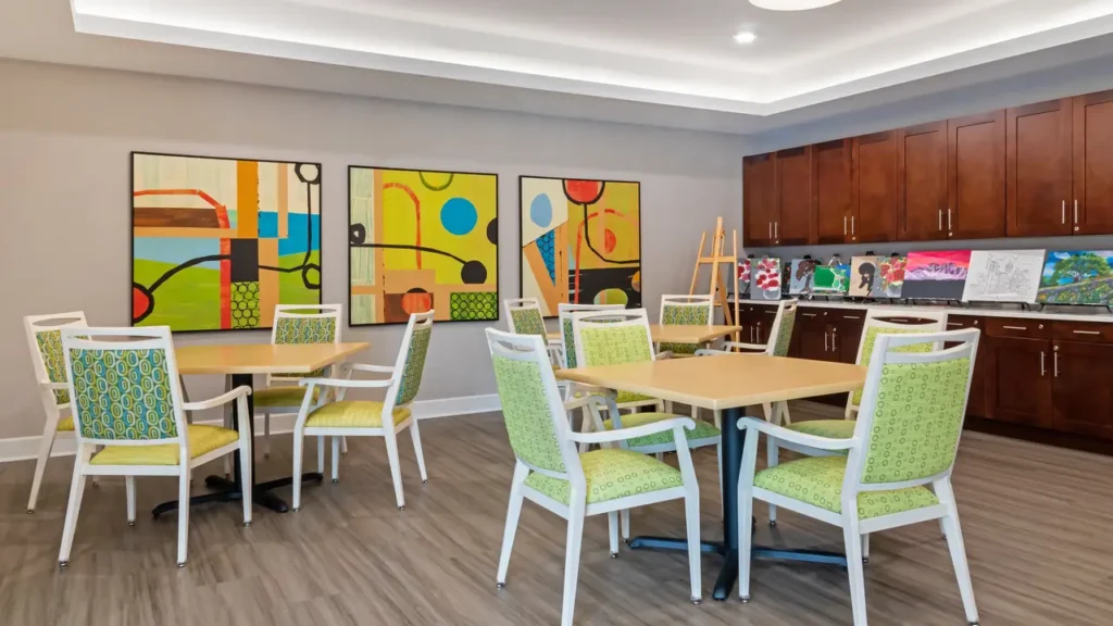 A brightly lit room with modern decor, featuring tables and chairs with green patterned upholstery. The walls display abstract paintings, and cabinets hold various supplies and artwork. The floor is covered with light wood flooring.