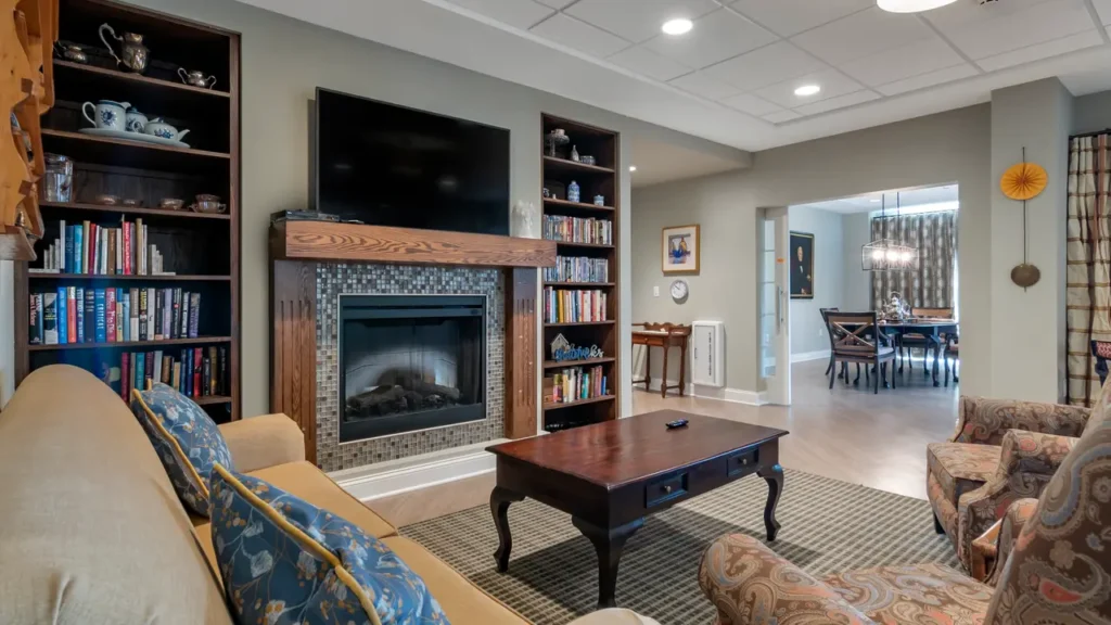 A cozy living room with beige walls features a fireplace with a TV mounted above it, flanked by wooden bookshelves filled with books and decorative items. A wooden coffee table sits in front of a beige couch and patterned armchair. An adjacent dining area is visible.