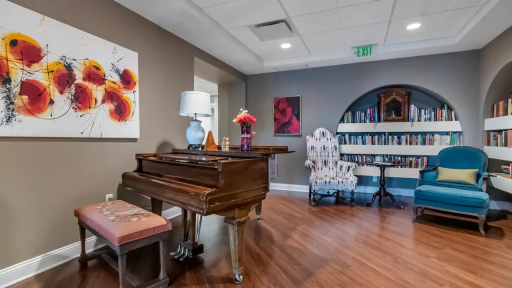 A cozy room with wooden flooring features a grand piano with a cushioned stool, a floral armchair, a blue chair, and a bookshelf filled with books. Art pieces and framed photos adorn the walls, and a table with flowers and a lamp sits beside the piano.