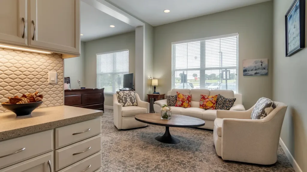 A modern living room with light green walls featuring a white sofa adorned with colorful cushions, two white armchairs, a round coffee table, and a TV on a wooden stand. The room is well-lit with natural light from two large windows. Kitchen area visible on the left.