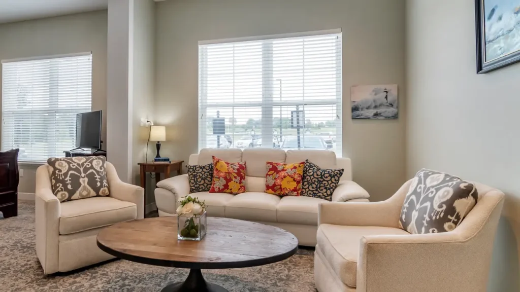 A cozy living room with a white sofa adorned with colorful pillows, flanked by two beige armchairs. A wooden coffee table with a floral centerpiece sits in the center. The room is well-lit by natural light from a large window behind the sofa.