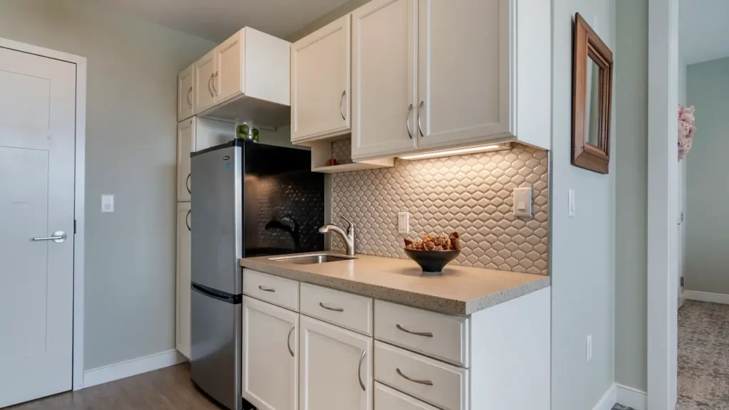 Modern kitchen with white cabinets, stainless steel refrigerator, and a light gray countertop. Under-cabinet lighting illuminates a textured backsplash with a geometric pattern. A small bowl of fruit sits beside the sink. Door and hall entrance visible.
