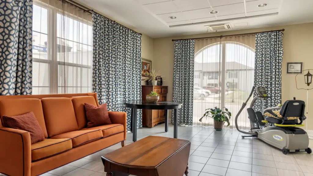 A cozy room with large windows covered by geometric-patterned and sheer curtains. An orange sofa with two striped cushions sits next to a coffee table. A round table with plants and decor is in the corner, and an exercise bike is near the window.